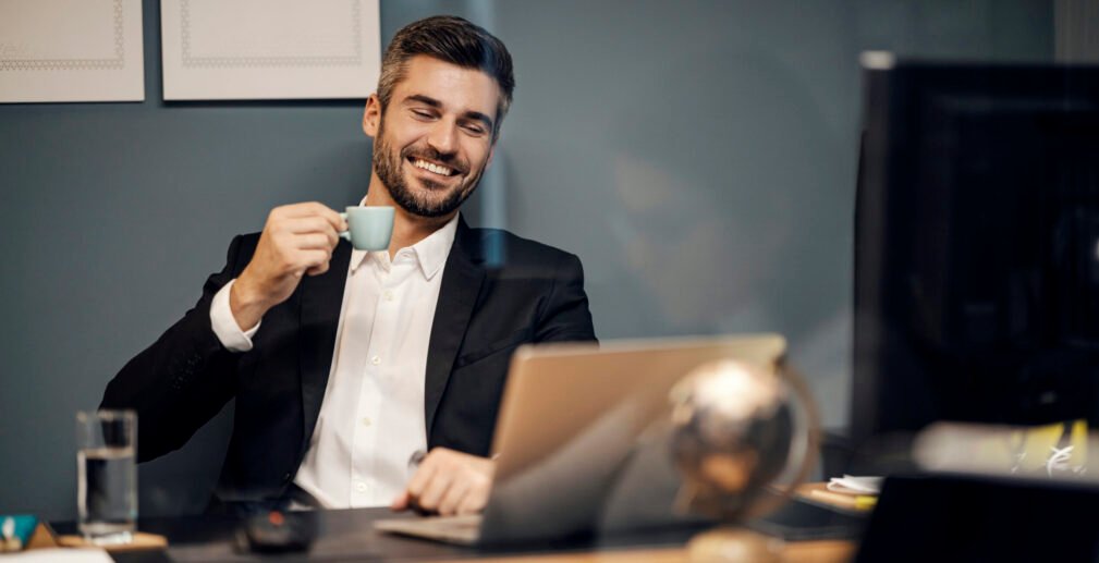 A cheerful executive sitting in his office and drinking coffee while smiling at the laptop.