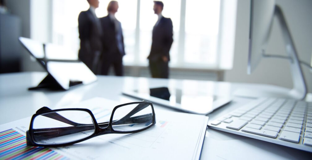 Technological devices, eyeglasses and financial document at workplace on background of three businessmen discussing ideas